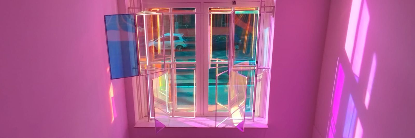 Tamar Frank, Reflecting Angles: hinged acrylic panels with dichroic film and LED-lit edges mounted in the tall window of 30m3__, casting pink, violet and blue reflections across the walls, with a parked car visible in the street beyond